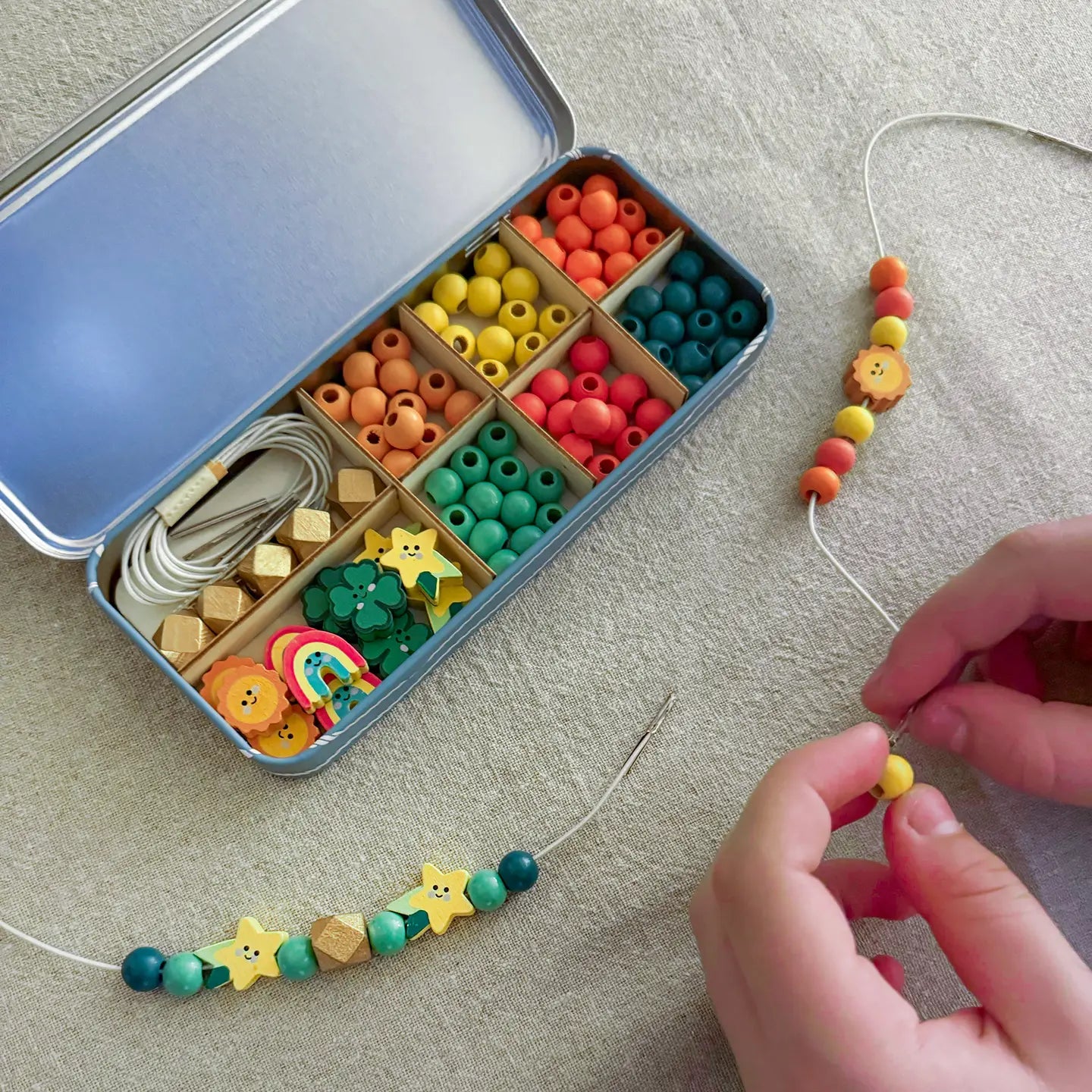 Colorful bead set in a tin with hands stringing beads on a gray surface