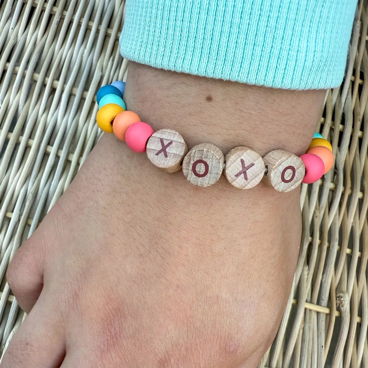 Colorful beaded bracelet with wooden beads displaying 'XO' on a wrist against a woven background.